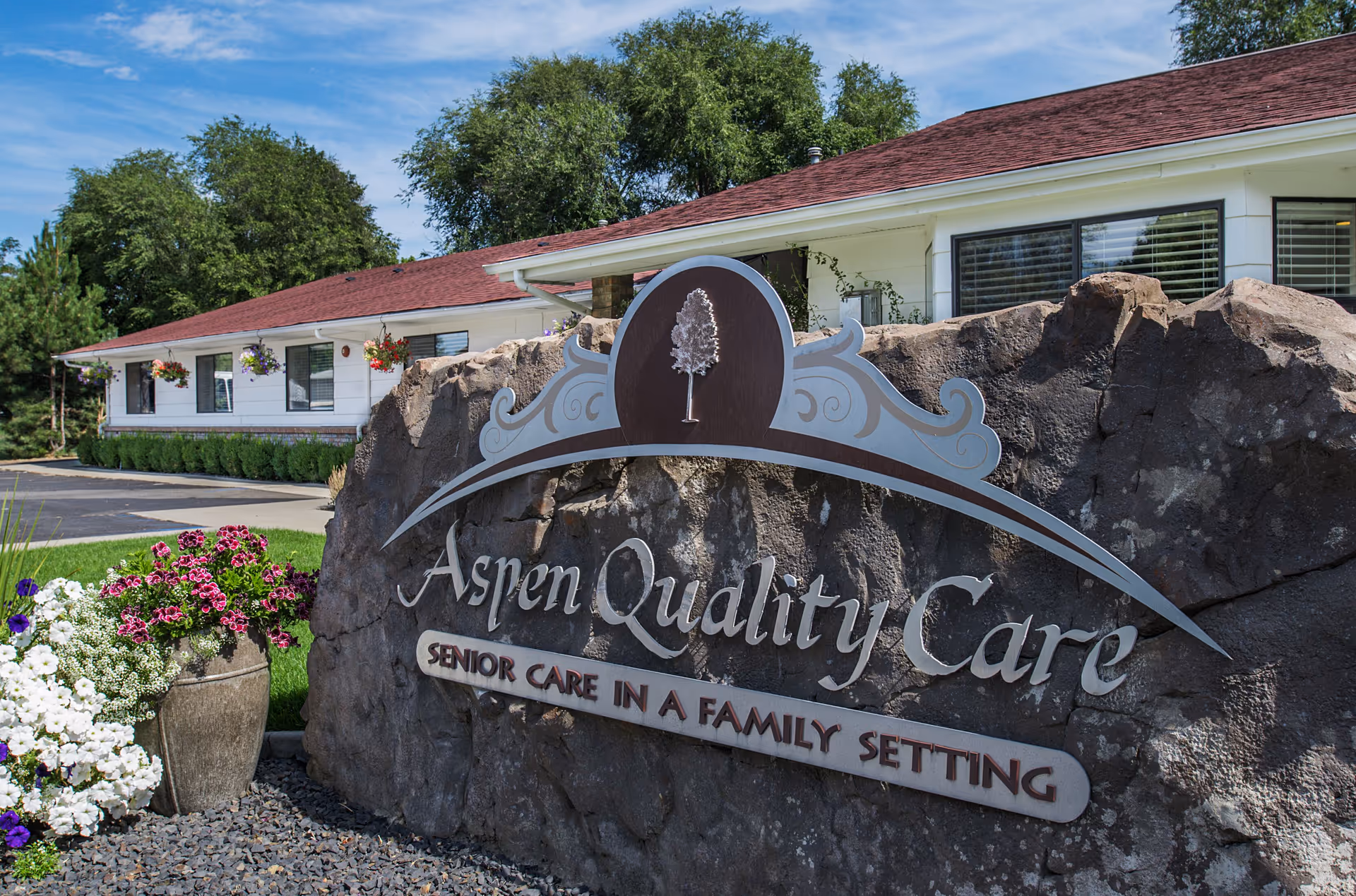 Outdoor view of Aspen Quality Care Inc. facility sign mounted on a large rock with flowers planted nearby and a white building with a red roof and hanging flower baskets in the background under a blue sky.