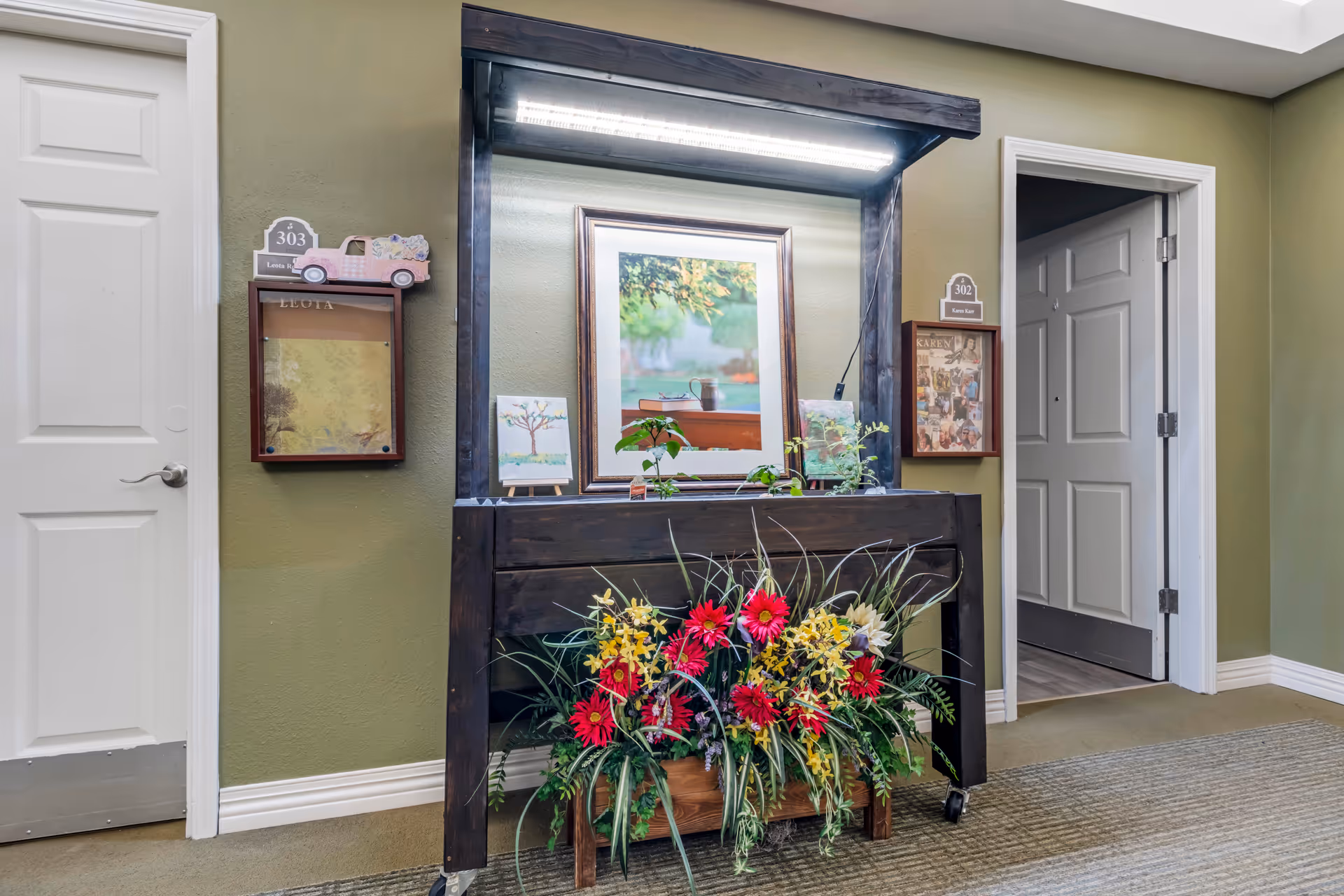 Interior hallway of a senior living facility with two white doors labeled 302 and 303. Between the doors is a dark wooden planter with colorful artificial flowers and small potted plants on top. Above the planter is a framed picture of a cup and books on a table, illuminated by a light fixture. The walls are painted green and the floor is carpeted.