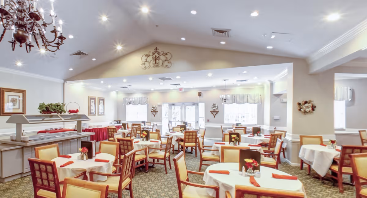 Bright, spacious dining room with round tables set with white linens and red napkins, wooden chairs, chandeliers and a buffet service area.