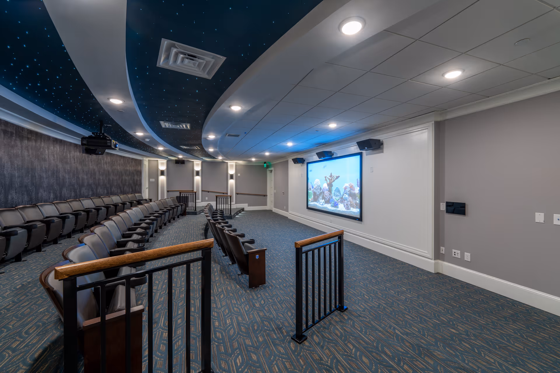 Interior view of a small theater room with rows of cushioned seats facing a large screen displaying an underwater scene. The ceiling has a curved design with star-like lights, and the walls are painted in neutral tones with recessed lighting.