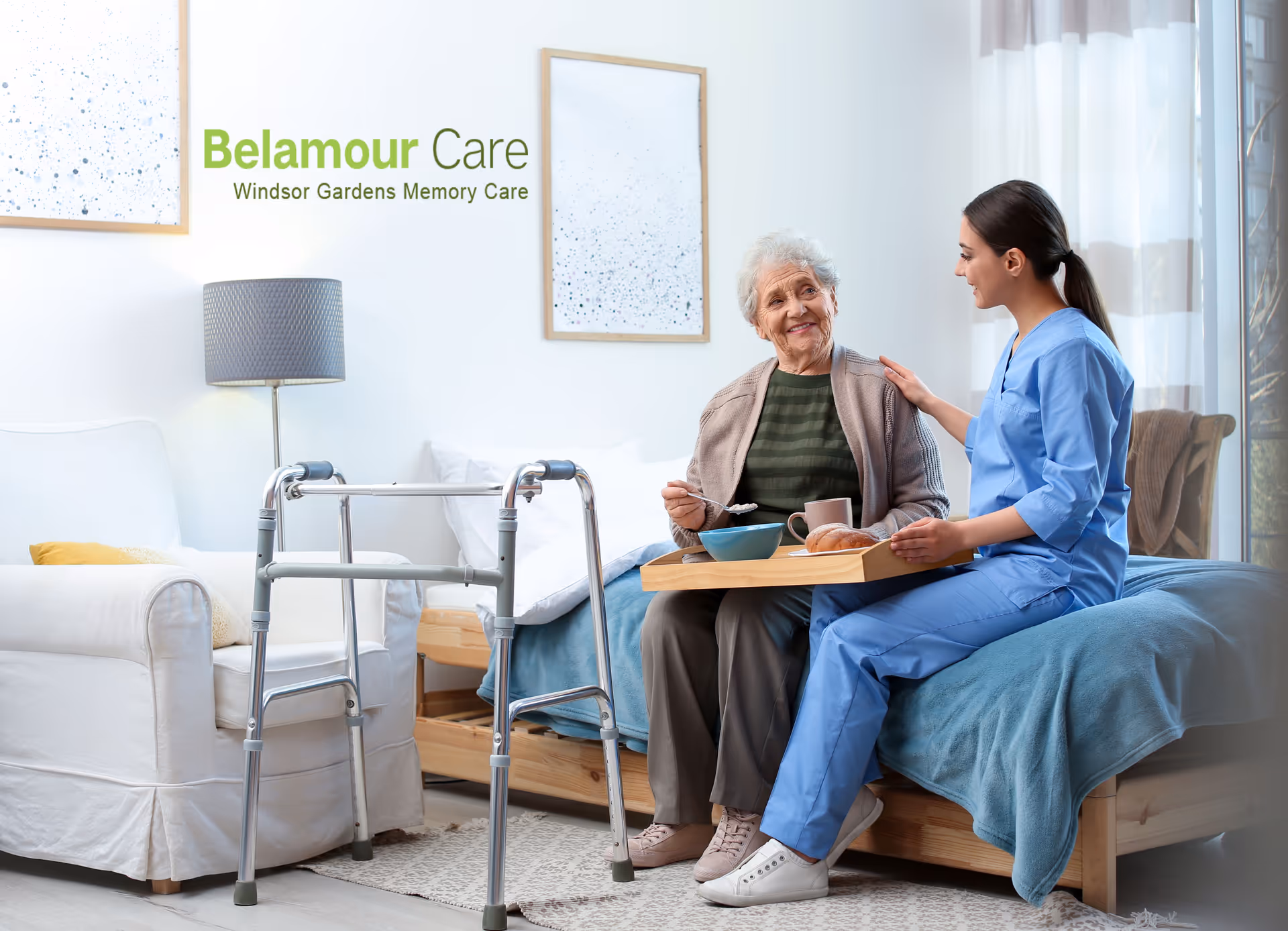 A caregiver in scrubs sits on a bed with an elderly woman, offering a breakfast tray in a bright memory care room with a walker nearby.
