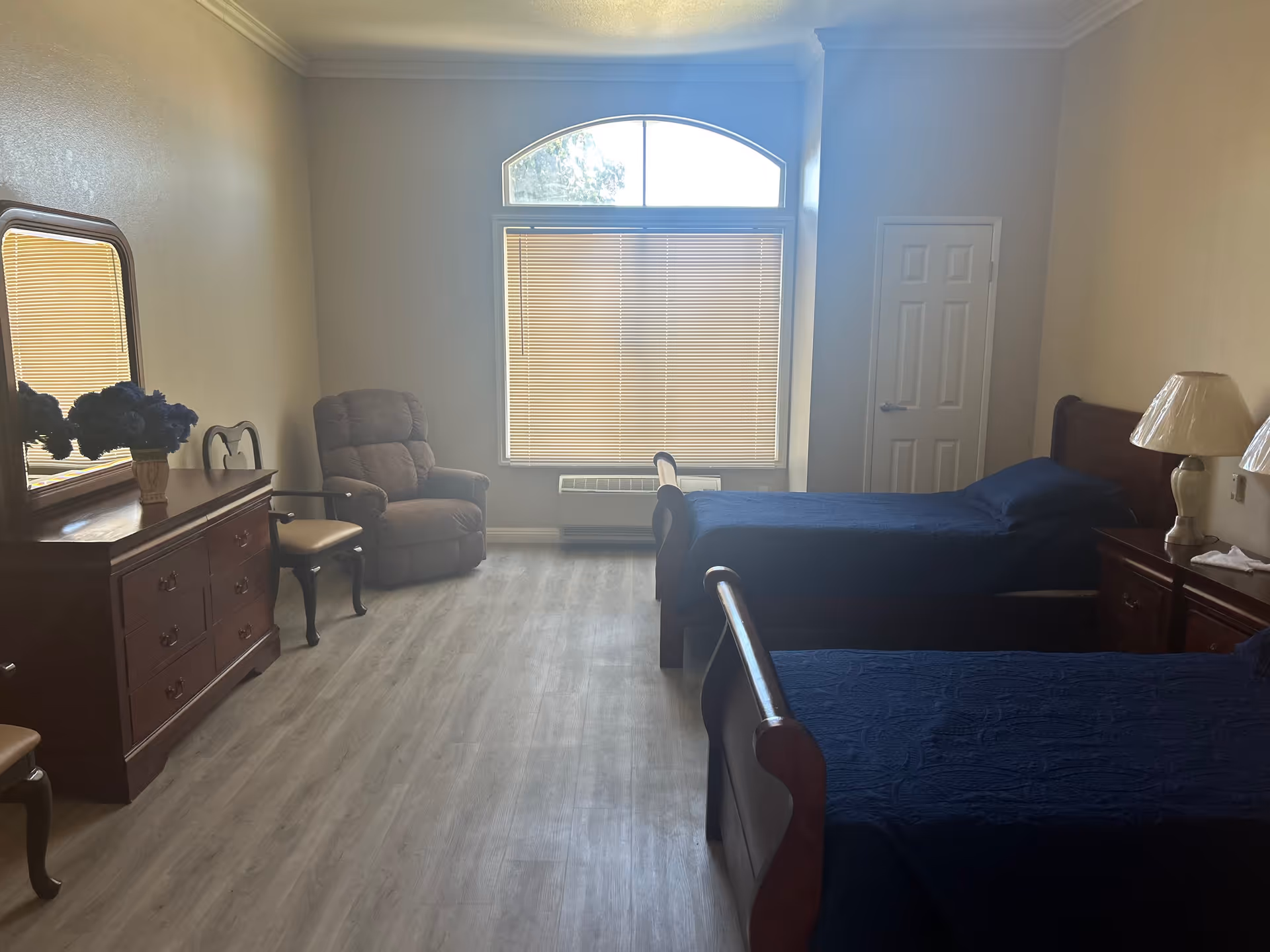 A bedroom with two wooden twin beds covered in dark blue bedspreads. There is a wooden dresser with a mirror and a vase of blue flowers on top. Two chairs and a brown recliner are positioned near a large window with blinds. A nightstand with a lamp is next to one of the beds. The room has light-colored walls and wood flooring.