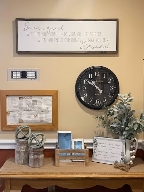 A beige wall with a decorative sign that reads 'Be our guest whether you come in to visit, or just to rest when you enter our home may you be blessed.' Below the sign is a black wall clock showing the time as 10:50. On a wooden table beneath the clock are decorative items including two wooden jars with spherical ornaments, a wooden holder with cards, a framed document, and a plant with green leaves and white beads.