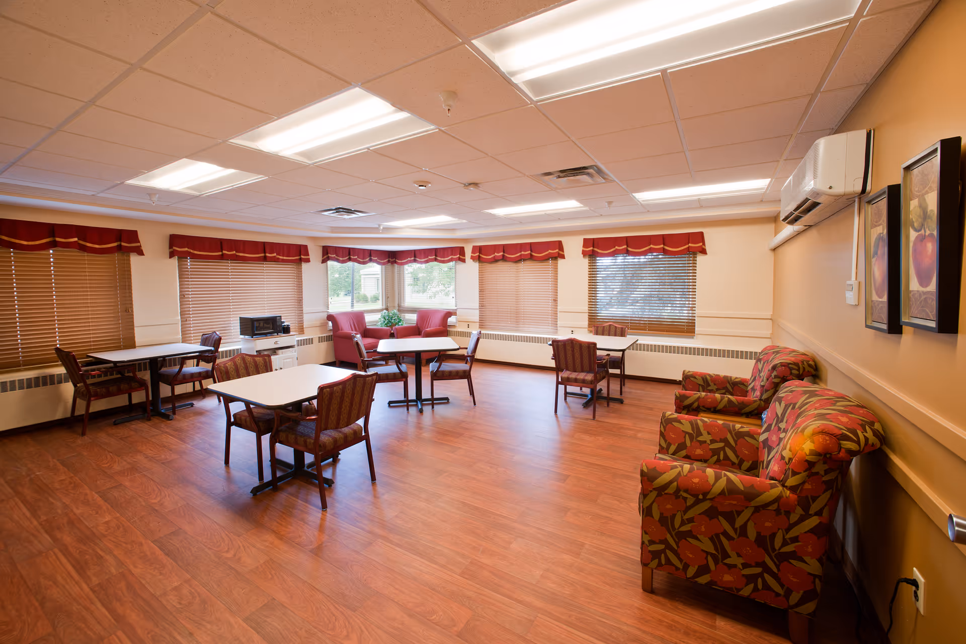 A well-lit room with wooden flooring and several tables with chairs arranged around them. The windows have wooden blinds with red valances. There are two floral-patterned armchairs along the right wall beneath two framed pictures. A microwave is placed on a small white cabinet near the windows. The ceiling has recessed lighting and an air conditioning unit is mounted on the right wall.