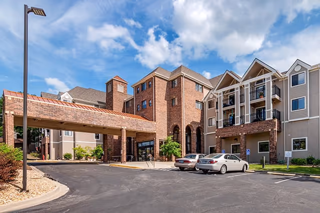 Exterior view of Brookdale Wornall Place senior living facility showing a multi-story building with brick and beige siding, a covered entrance, parked cars, and a clear blue sky with some clouds.