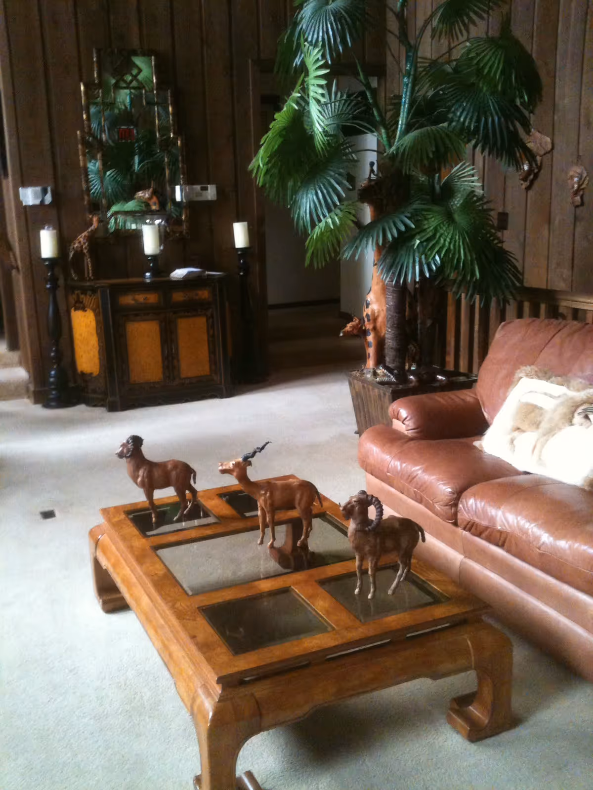 A cozy living room with a brown leather sofa adorned with a light-colored throw pillow and blanket. In front of the sofa is a wooden coffee table with glass insets, displaying three decorative animal figurines. To the right of the sofa is a large potted artificial palm tree. In the background, there is a wooden cabinet with three candles on tall candle holders and a mirror above it, reflecting part of the room. The walls are wood-paneled, and the floor is carpeted in a light color.
