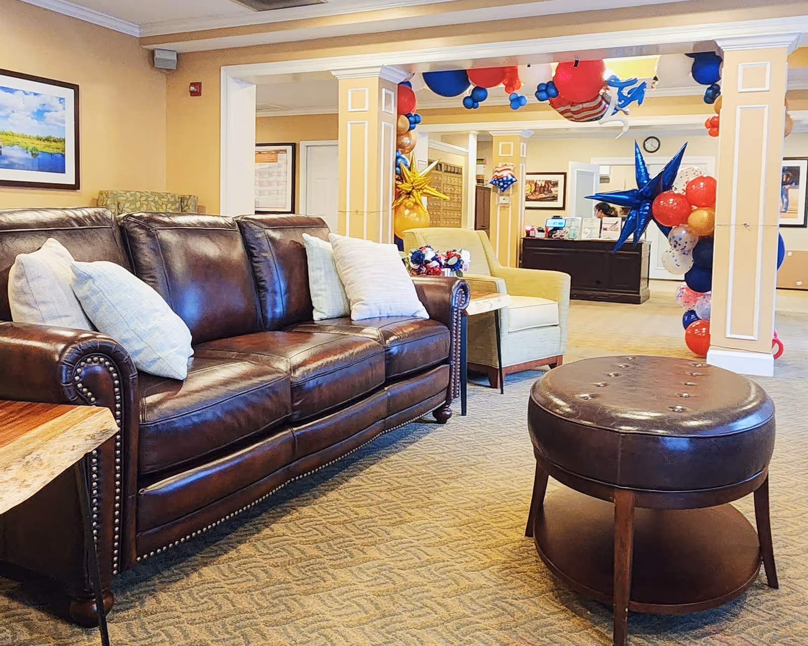 A cozy living room area in a senior living facility featuring a dark brown leather sofa with light-colored pillows, a round leather ottoman, and a beige armchair. The room is decorated with patriotic red, white, and blue balloons and star-shaped decorations. In the background, there is a reception desk with a person seated behind it and framed artwork on the walls.
