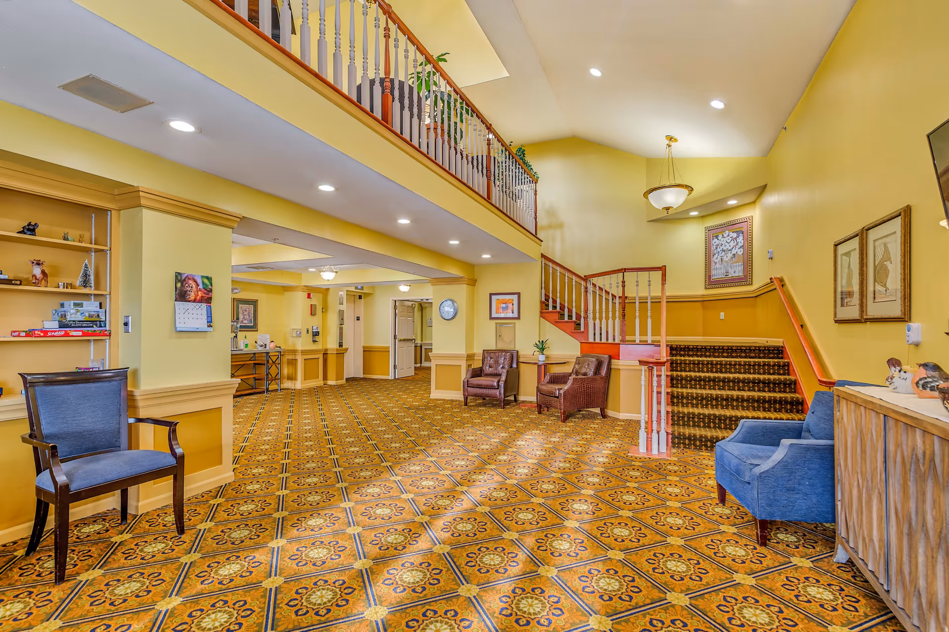 Interior view of a senior living facility lobby area with yellow walls and patterned carpet. The space features a staircase with wooden railings leading to an upper floor, several chairs including a blue armchair and two brown chairs, framed artwork on the walls, and a wooden cabinet with decorative items. The ceiling has recessed lighting and a hanging light fixture.