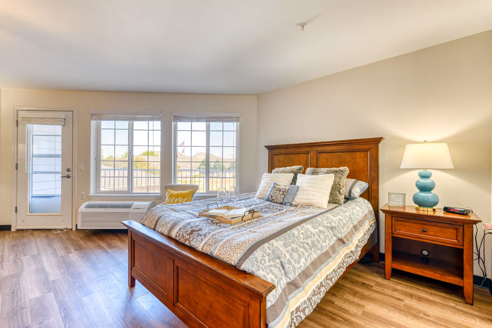 Well-lit bedroom with a wooden bed, nightstand and lamp, wood flooring, and large windows with a door to the outside.