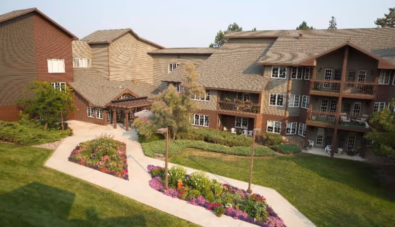 Exterior view of a multi-story senior living facility with brown and beige siding, surrounded by well-maintained green lawns and colorful flower beds along a paved walkway.