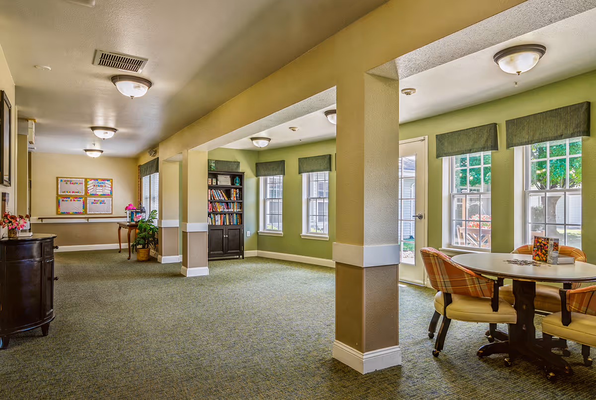 Communal lounge with a round table and chairs, bookshelf, large windows, and carpeted flooring.