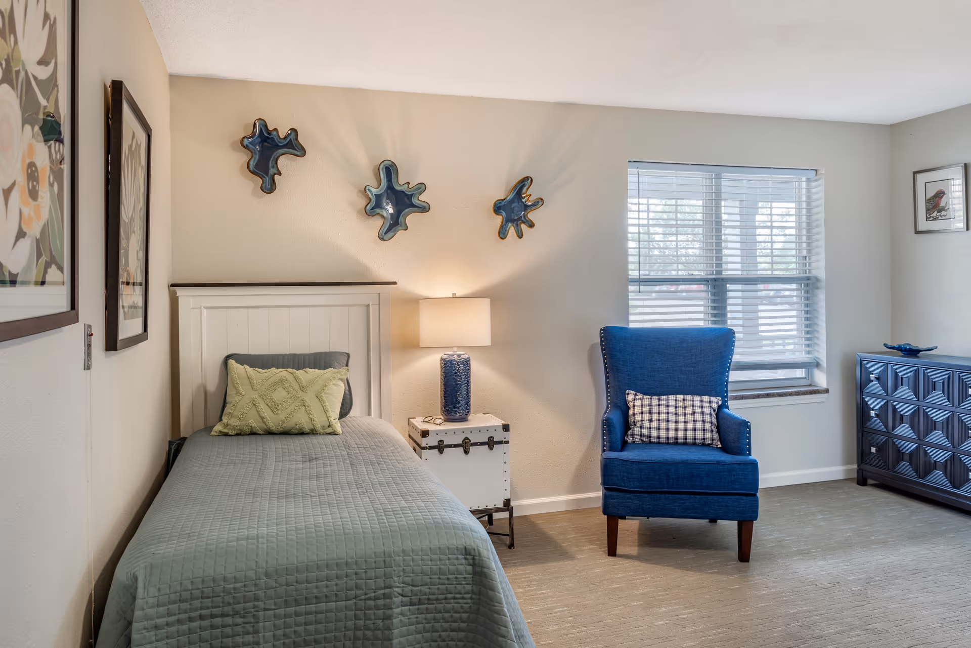 A neatly arranged bedroom with a single bed covered in a green quilt and a green decorative pillow. Next to the bed is a white nightstand with a blue lamp. On the wall above the bed are three blue abstract wall decorations. To the right is a blue armchair with a checkered pillow, positioned near a window with white blinds. A blue dresser with a decorative bowl on top is against the far wall. The room has light-colored walls and carpeted flooring.