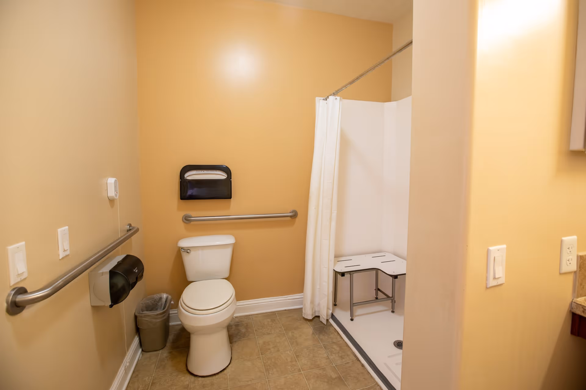 A senior living facility bathroom with beige walls and tiled floor. The room features a white toilet with grab bars on the walls for accessibility, a small trash can, a toilet paper dispenser, and a wall-mounted paper towel dispenser. To the right, there is a walk-in shower with a white curtain and a white shower bench inside.
