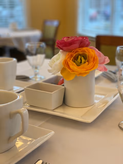 Close-up of a dining table set with white cups, a small white rectangular dish, and a white vase holding colorful artificial flowers in shades of pink, orange, and white. The background shows blurred chairs and windows.