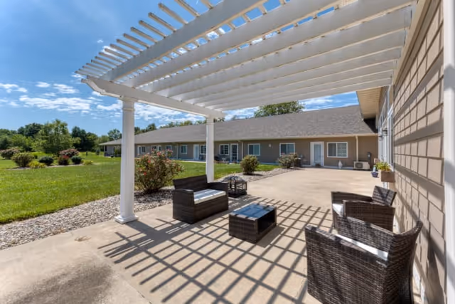 Outdoor patio area with a white pergola casting shadows on the concrete floor. The patio is furnished with dark wicker chairs and a small table. In the background, there is a single-story building with multiple windows and doors, surrounded by green grass and bushes under a blue sky with some clouds.