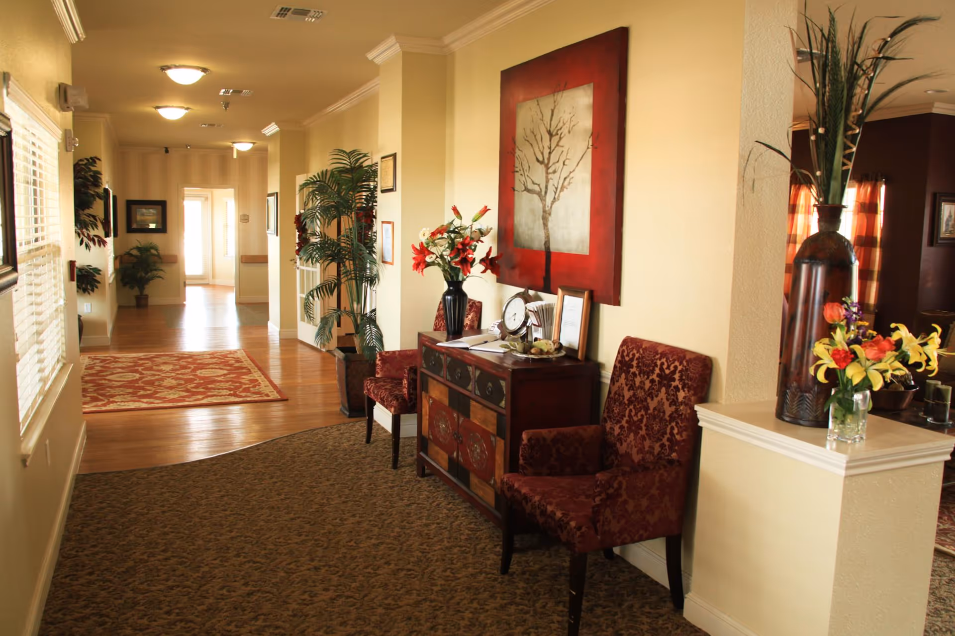 Well-decorated interior hallway/lounge with patterned chairs, a console table topped with flowers and artwork, potted plants, and a mix of carpeted and hardwood flooring.