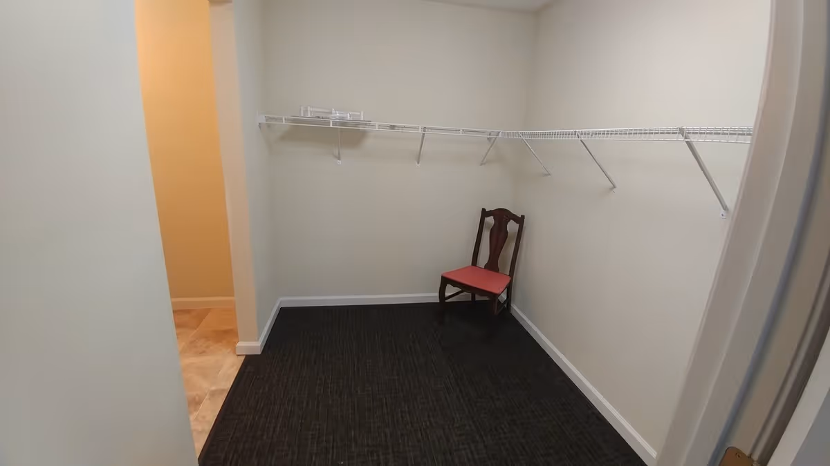 Empty walk-in closet with white wire shelving along two walls and a single wooden chair with a red cushion placed against the far wall. The closet has beige walls and dark carpet flooring, with an open doorway leading to a room with tiled flooring.