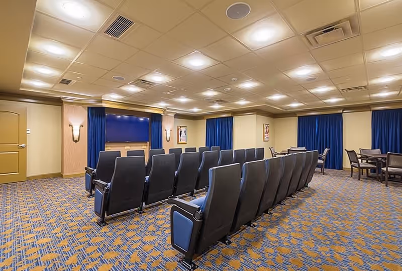 A theater-style room with rows of black cushioned chairs facing a large flat-screen TV mounted on the wall. The room has patterned carpet, beige walls with blue curtains covering the windows, and several small tables with chairs in the back corner.