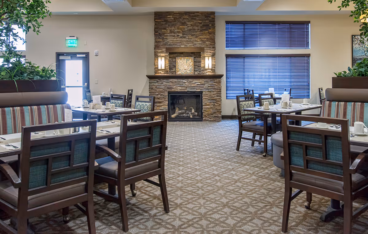 Dining area in an assisted living community with multiple tables and chairs arranged neatly. The room features a stone fireplace in the center, large windows with blinds on the right, and potted plants on the left. The tables are set with white cups, plates, and utensils, ready for a meal.