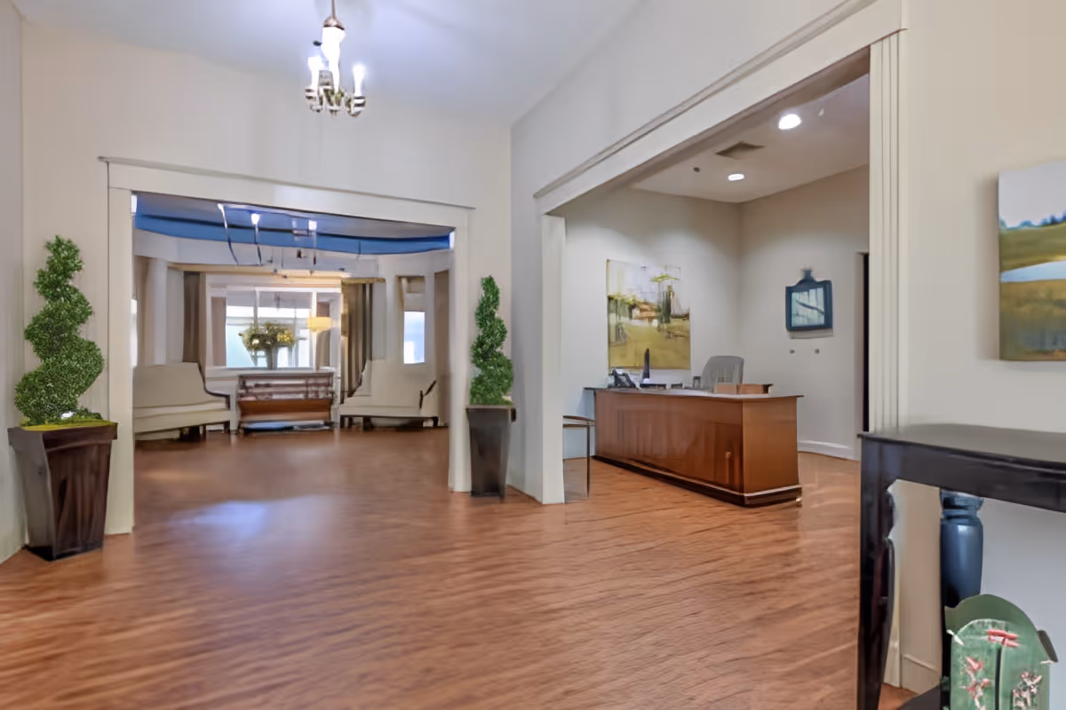 Interior view of a senior living facility lobby area with wooden flooring, a reception desk on the right, decorative plants in tall planters, seating area with benches and a table with flowers in the background, and artwork on the walls.