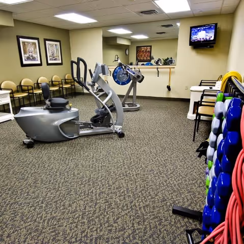 A fitness room in a senior living facility with exercise equipment including a rowing machine and an elliptical trainer. There are chairs lined up against the wall, framed artwork, a wall-mounted TV, and a rack holding colorful exercise balls and resistance bands.