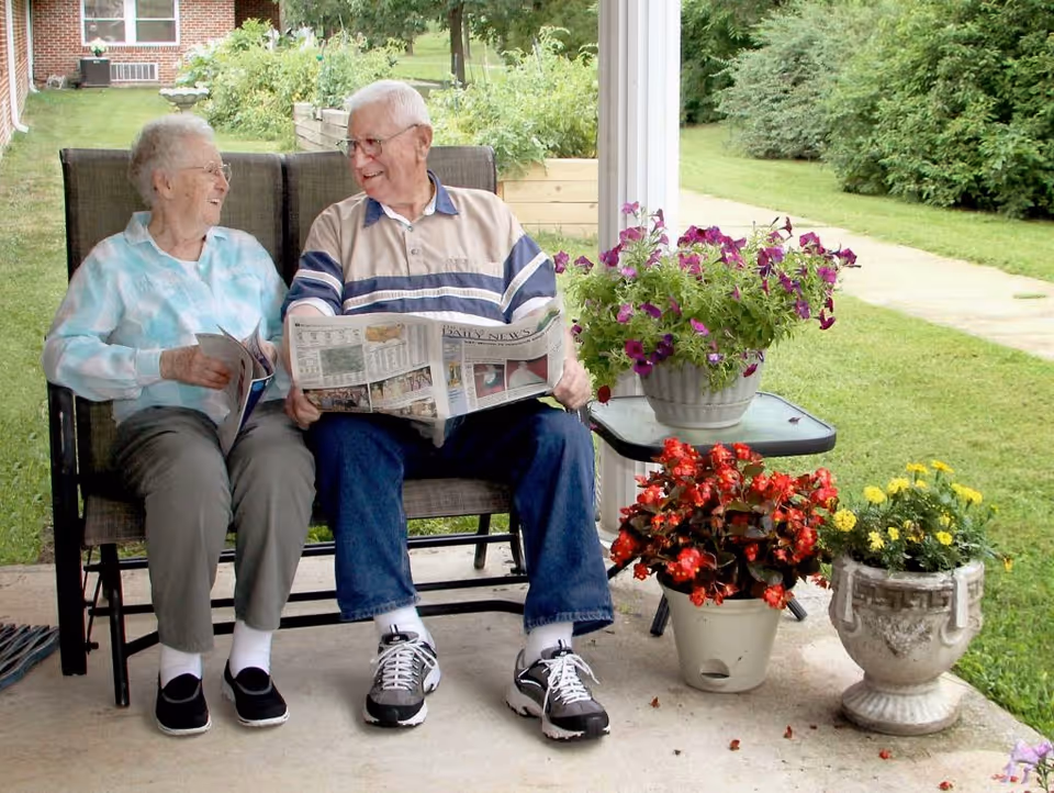 An elderly man and woman sitting on a bench on a covered patio, smiling and looking at each other. The man is holding a newspaper, and there are three flower pots with colorful flowers on the ground and a small table beside them. Green grass and bushes are visible in the background.
