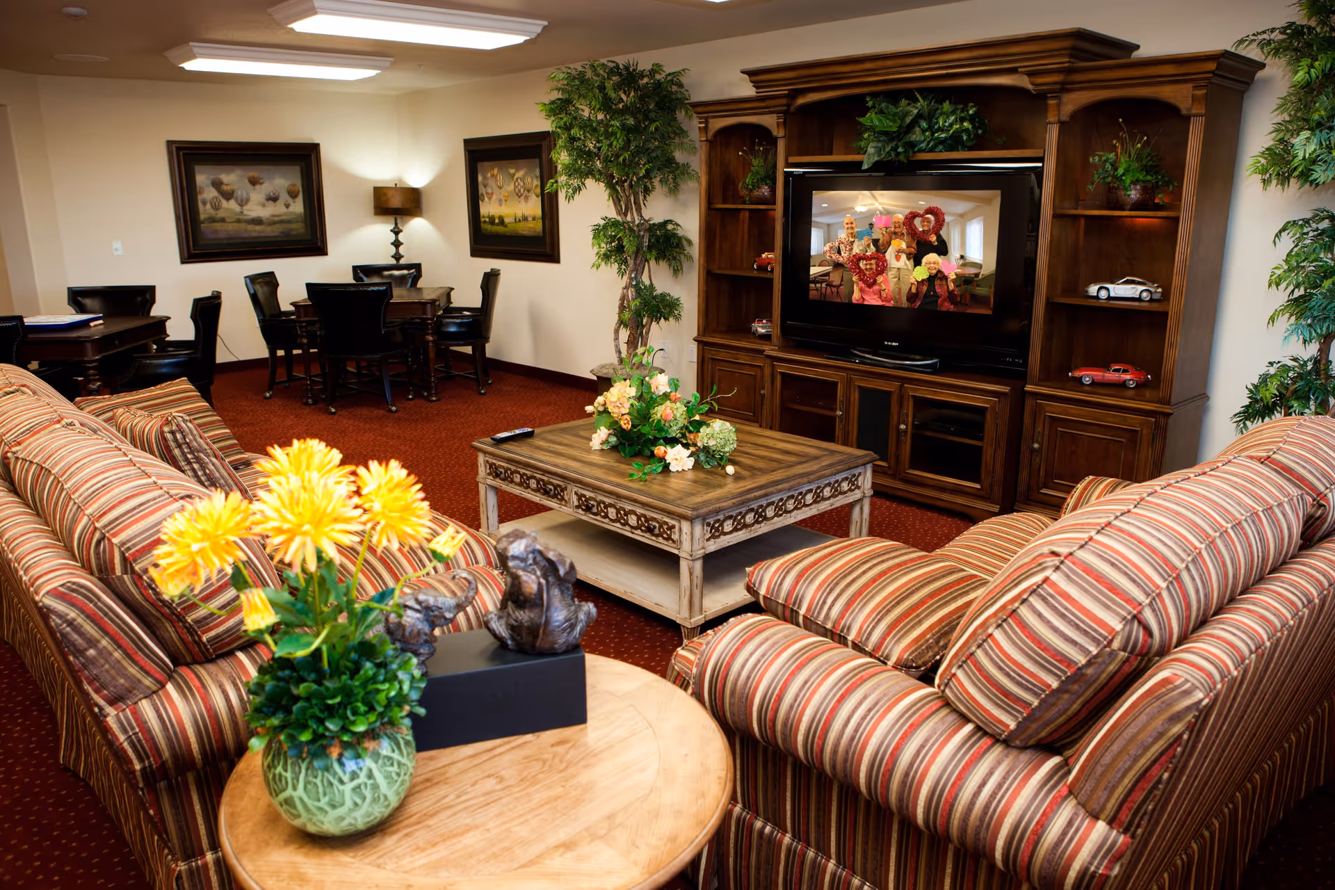 A cozy living room area in a senior living facility with two striped sofas facing a wooden entertainment center with a TV. The TV displays a group of elderly people holding heart-shaped decorations. The room has a red carpet, a wooden coffee table with a flower arrangement, side tables with plants, and framed artwork on the walls. There is also a small dining area with a table and chairs in the background.