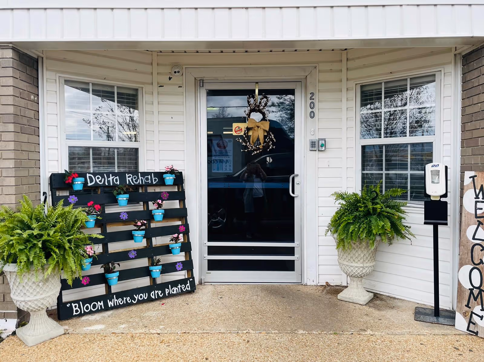 Entrance to Delta Rehabilitation and Healthcare Center of Cleveland with a glass door decorated with a wreath. On the left side, there is a black wooden pallet with small blue pots containing flowers and the words 'Delta Rehab' and 'Bloom where you are planted' written on it. Two large potted ferns flank the entrance. A hand sanitizer dispenser is mounted on a stand to the right of the door. The building exterior features white siding and brick walls.