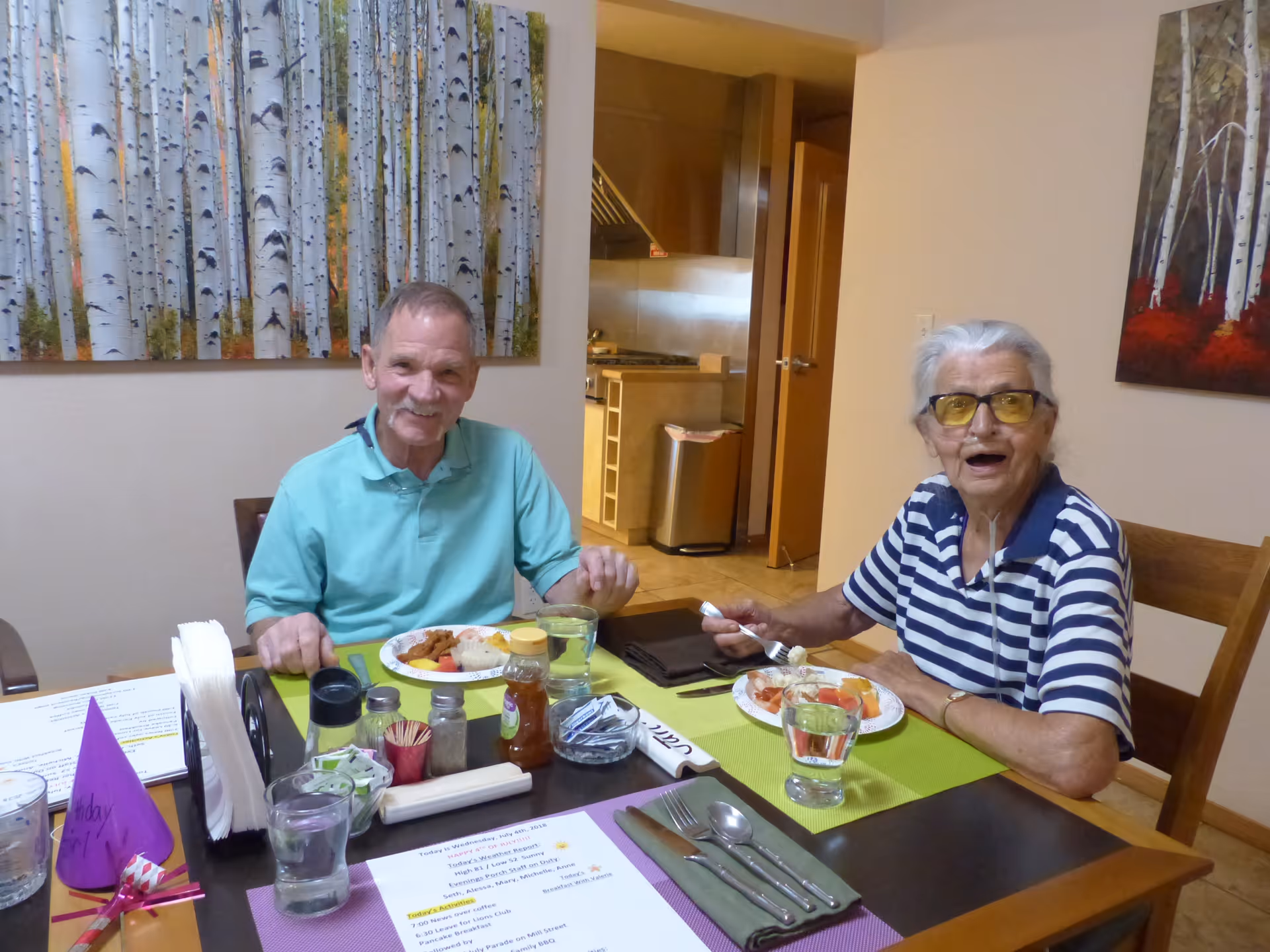 Two elderly people sitting at a dining table in a well-lit room, enjoying a meal together. The man on the left is wearing a light blue polo shirt and smiling, while the woman on the right is wearing a striped shirt and yellow-tinted glasses, holding a fork with food. The table is set with plates of food, glasses of water, condiments, utensils, and a printed menu. Behind them, there are paintings of birch trees on the walls and a view into a kitchen area.