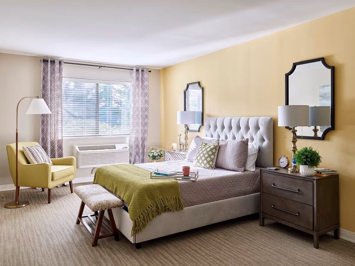 A bright and cozy bedroom with a large window covered by patterned curtains. The room features a tufted upholstered bed with multiple pillows and a green throw blanket. There is a wooden bench at the foot of the bed, a yellow armchair with a floor lamp beside it, and a dark wooden nightstand with a lamp, clock, plant, and books. Two decorative mirrors hang on the yellow wall behind the bed.