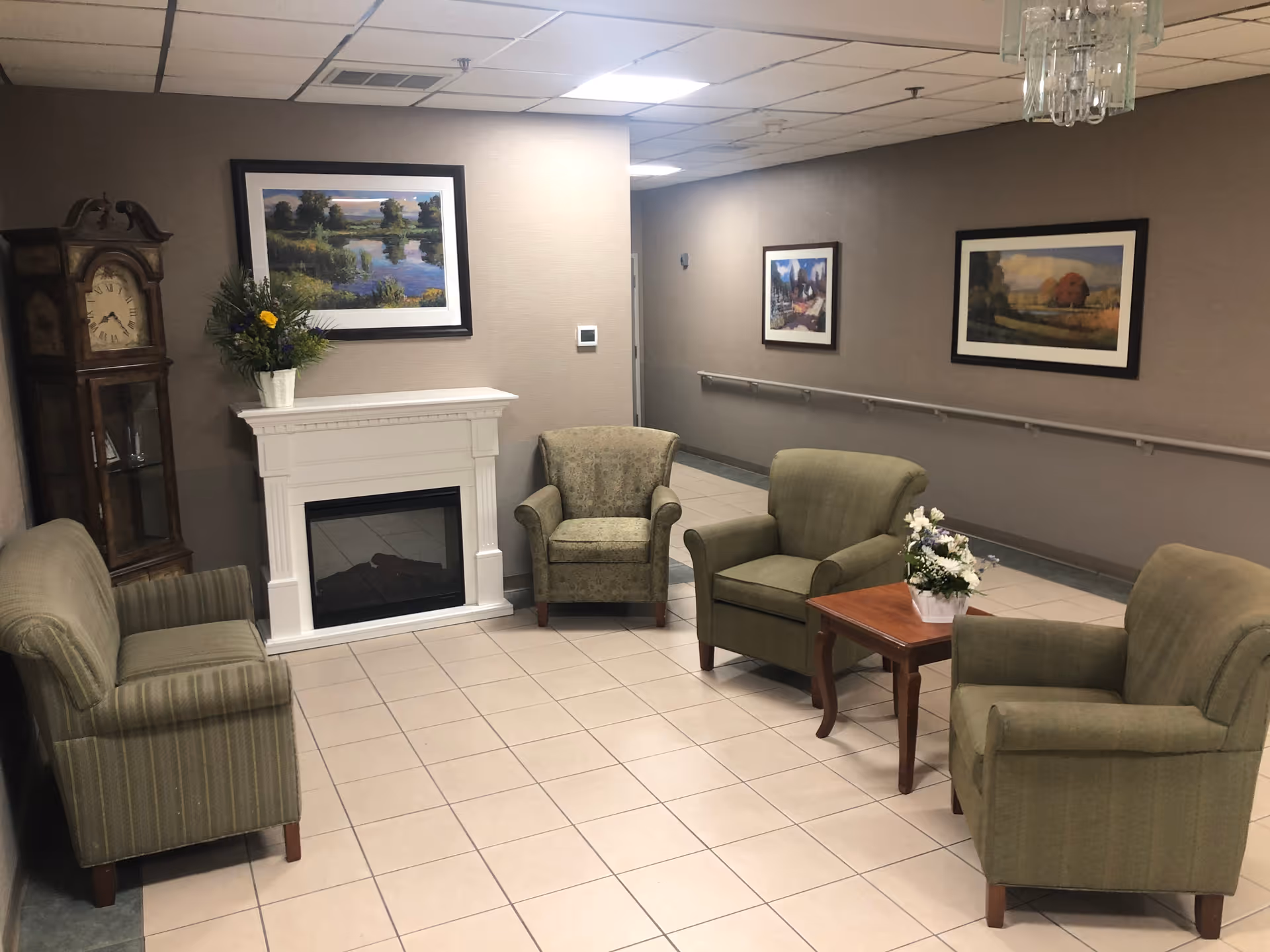 A cozy sitting area in a senior living facility with four green upholstered armchairs arranged around a small wooden table with a flower arrangement. There is a white electric fireplace against the wall, a tall grandfather clock to the left, and three framed landscape paintings on the walls. The floor is tiled, and the ceiling has recessed lighting.