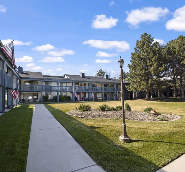 Outdoor view of a two-story retirement facility building with balconies and American flags displayed. There is a paved walkway leading through a grassy courtyard with a lamp post and some small shrubs. The sky is blue with scattered clouds.