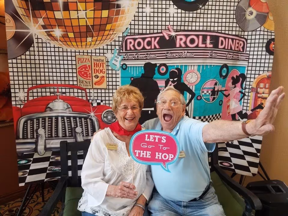 An elderly man and woman sitting on a bench in front of a colorful rock and roll diner-themed backdrop. The woman is smiling and wearing a white blouse with a red scarf, while the man is enthusiastically posing with his arm extended and holding a sign that says 'LET'S Go To THE HOP'. The backdrop features a large disco ball, a classic car front, vinyl records, and silhouettes of people dancing.