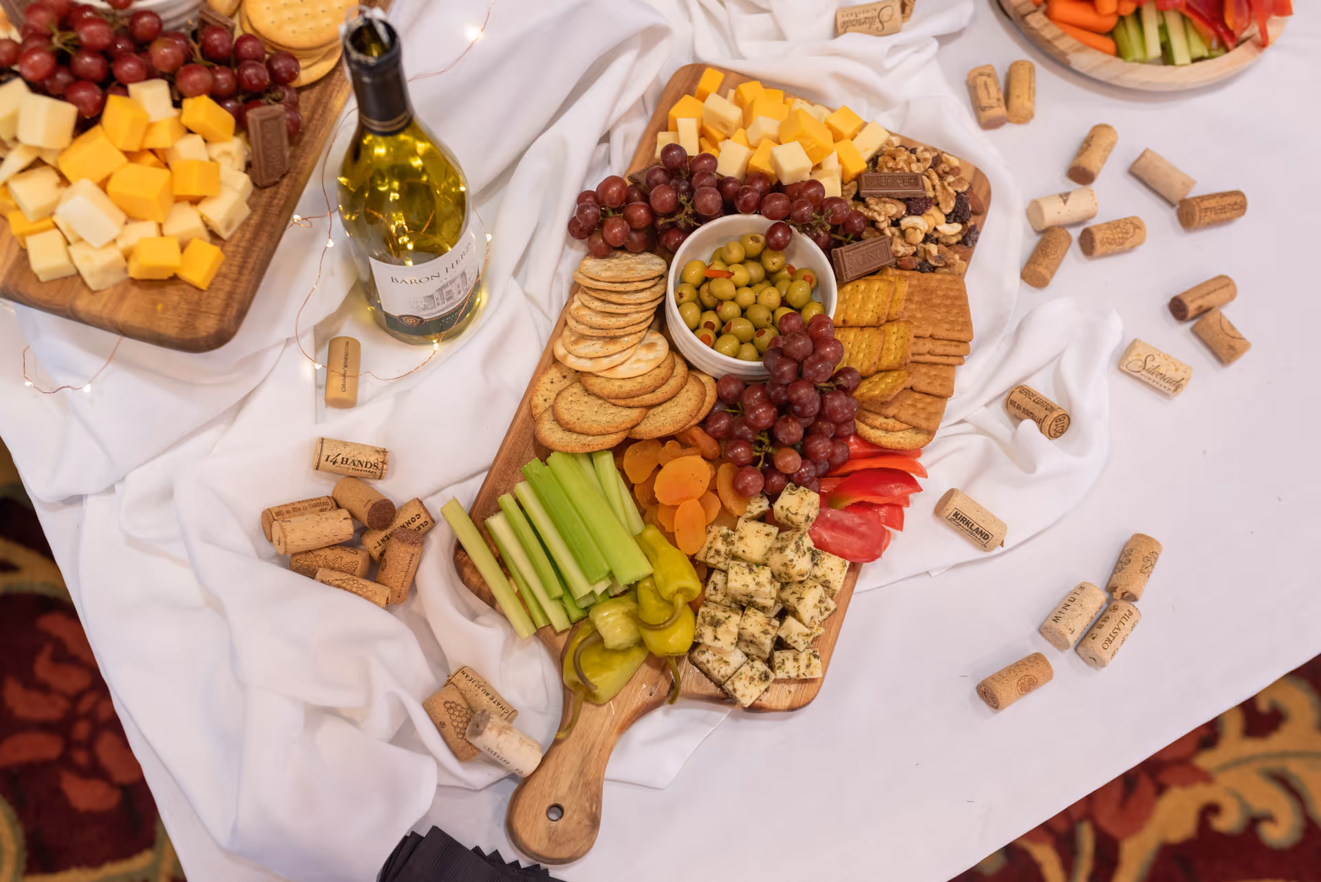 A wooden serving board with an assortment of snacks including cubed cheeses, red grapes, green olives in a bowl, crackers, celery sticks, pepperoncini, dried apricots, red bell pepper slices, herb-seasoned cheese cubes, and mixed nuts. A bottle of white wine and numerous wine corks are scattered on a white tablecloth underneath the board.