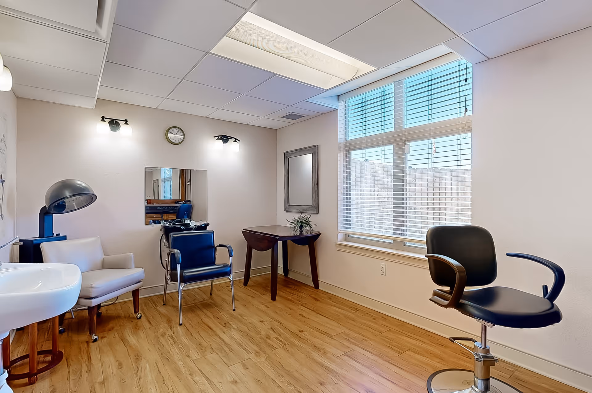 A small hair salon room with wooden flooring, a large window with blinds, a black salon chair, a white armchair, a hair dryer, a small table with a plant, a wall mirror, and two wall-mounted lights.