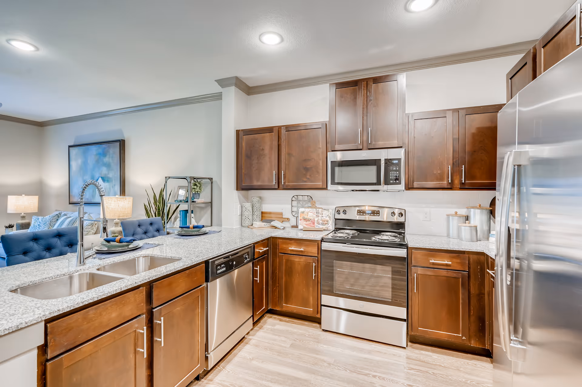 Modern kitchen with wooden cabinets, stainless steel appliances including a refrigerator, oven, microwave, and dishwasher. Granite countertops with a double sink and a high-arc faucet. Two blue cushioned chairs at the counter and a glimpse of a living room area with lamps and artwork in the background.