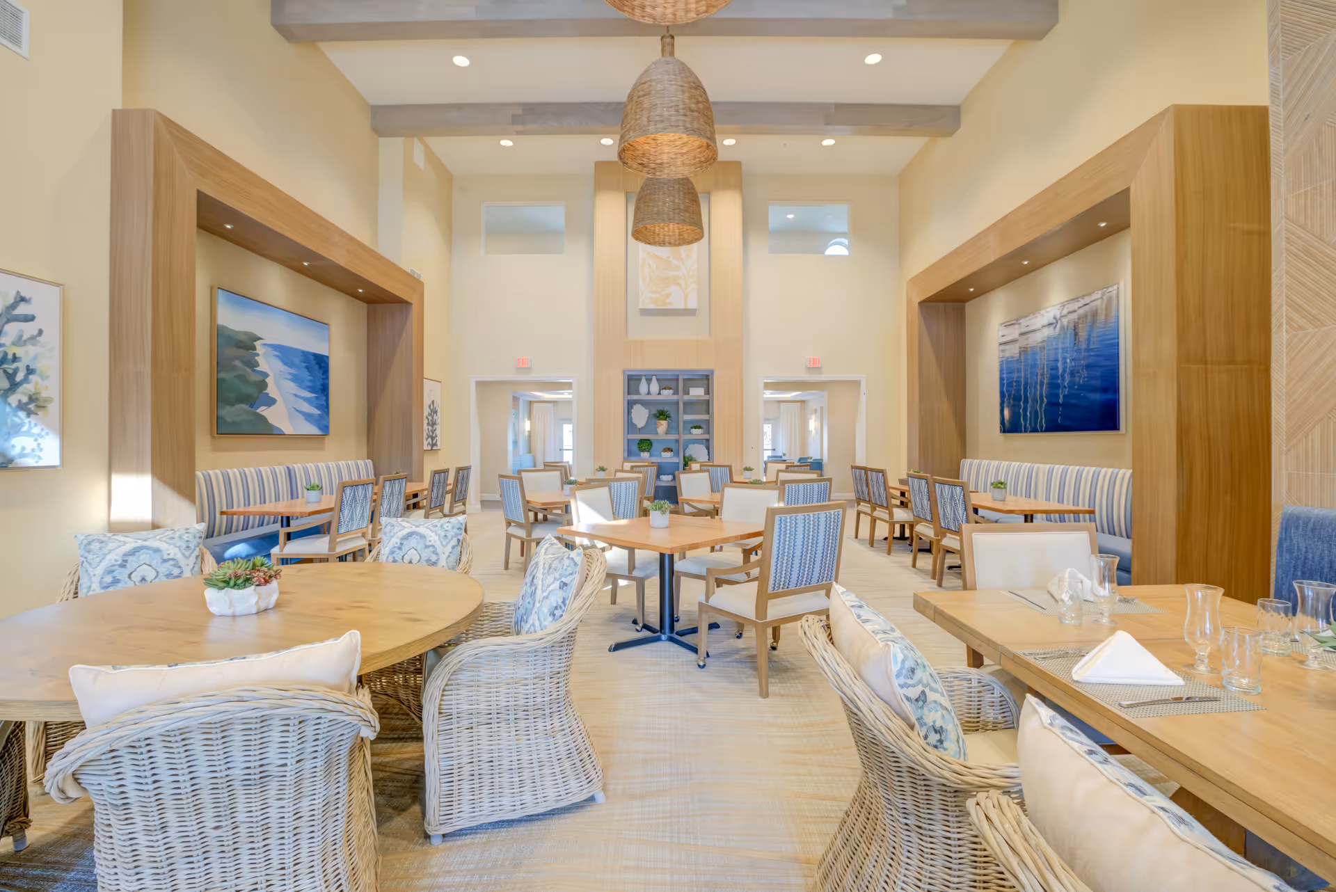 Bright, airy dining room in a senior living facility with multiple wooden tables, wicker chairs, banquette seating, and hanging woven pendant lights.