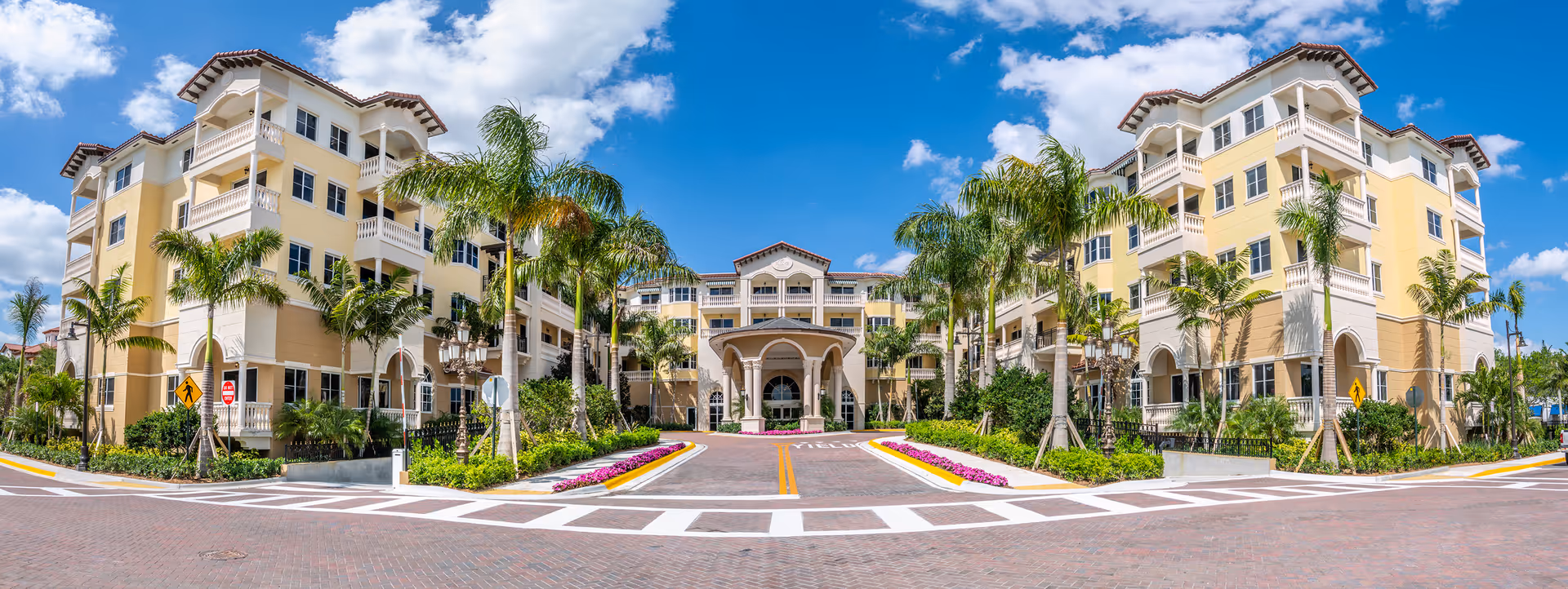 Front exterior view of The Palace at Weston, a large multi-story building with yellow and white walls, balconies, palm trees, and a covered entrance under a blue sky with scattered clouds.