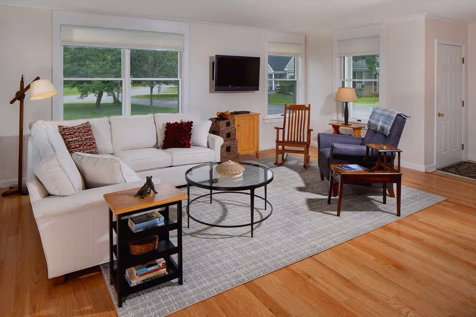 A bright and cozy living room with light-colored walls and hardwood floors. The room features a white sofa with decorative pillows, a wooden rocking chair, a blue armchair with a plaid throw, a round glass coffee table, and a small wooden side table with books. There is a floor lamp in the corner and a flat-screen TV mounted on the wall between two windows showing a green outdoor view.
