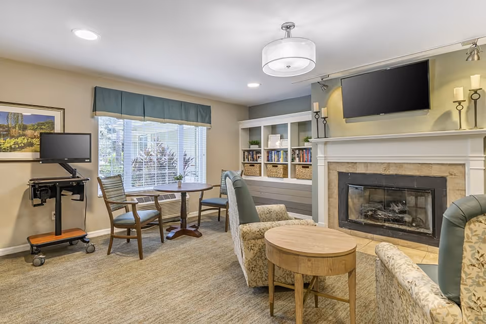 A cozy living room area in a senior living facility featuring a fireplace with a mounted flat-screen TV above it. There are two patterned armchairs and a small round wooden table in the center. A window with blinds and a blue valance lets in natural light. A small round table with two chairs is positioned near the window. A mobile computer workstation is placed against the wall. Built-in shelves with books and decorative baskets are visible next to the fireplace.