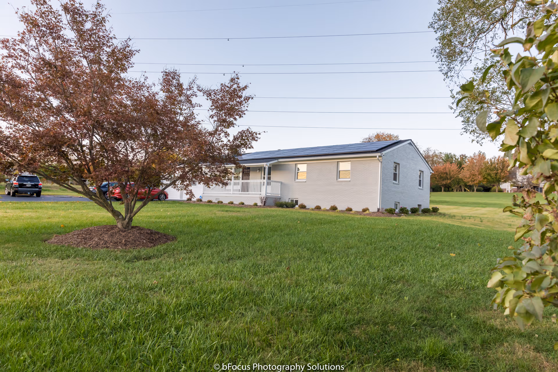 Single-story light-colored assisted living house on a grassy lawn with a tree and parked cars in the driveway.