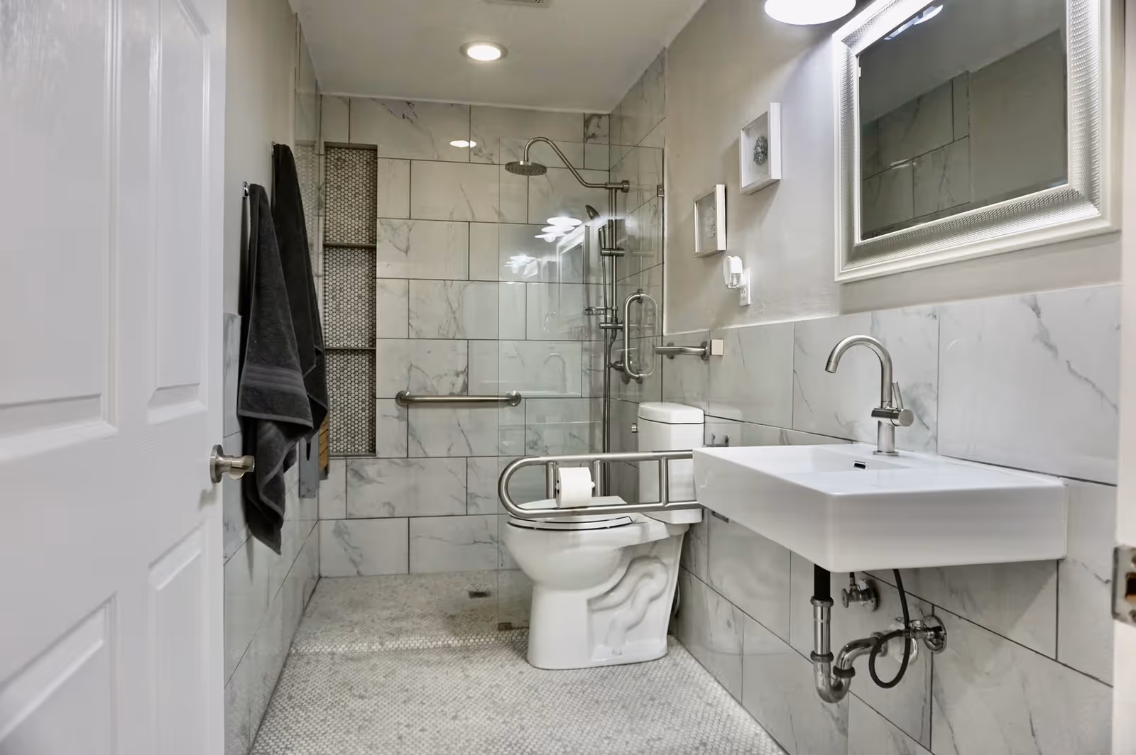 A modern bathroom with a walk-in shower featuring a glass door and a rainfall showerhead. The walls are tiled with large white marble tiles, and there is a white toilet with a safety grab bar and a mounted toilet paper holder. A white rectangular sink with a silver faucet is mounted on the wall, and a large mirror is above the sink. Two dark towels hang on a towel rack on the left wall. The floor has small mosaic tiles.