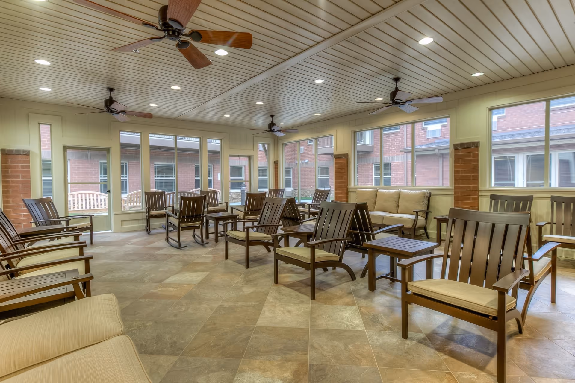 A spacious indoor seating area with multiple wooden chairs and small tables arranged neatly. The room has large windows on three sides allowing natural light to enter, ceiling fans, and a tiled floor. Outside the windows, a brick building and outdoor benches are visible.