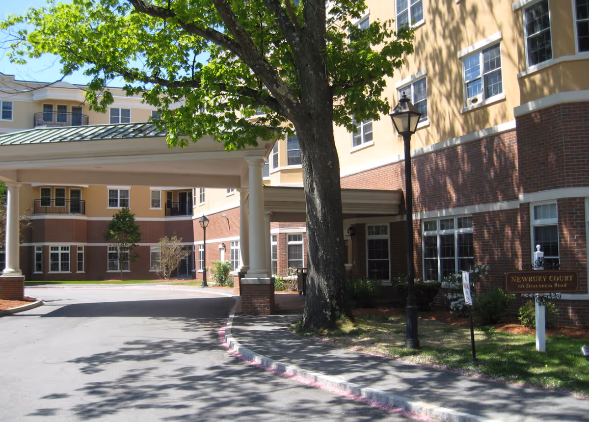 Front entrance of the Gardens at Newbury Court building with a porte-cochere, large tree, lamp posts, and a small sign on the lawn.