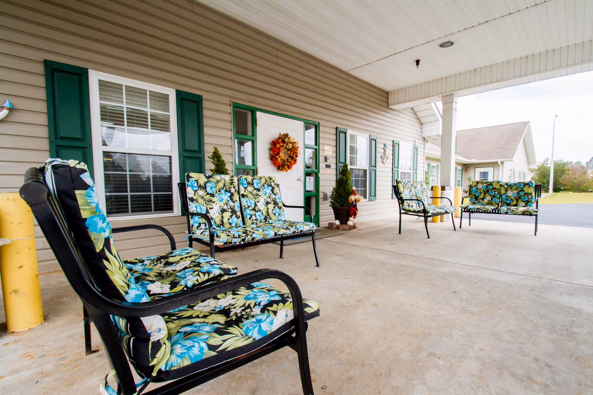 Covered outdoor seating area at Croatan Ridge Rehabilitation & Nursing Center with black metal chairs and benches featuring floral cushions. The building exterior has beige siding with green shutters and a white door decorated with a fall wreath. There are small potted plants near the entrance and a paved driveway visible in the background.