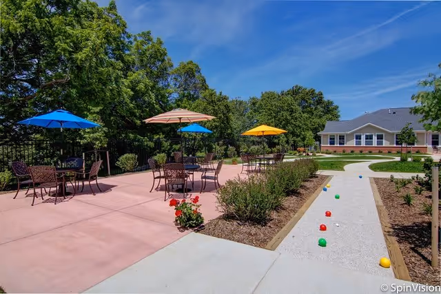 Outdoor patio area with several tables and chairs under colorful umbrellas, surrounded by greenery and trees. A bocce ball court with balls is visible on the right side, and a building is in the background under a clear blue sky.
