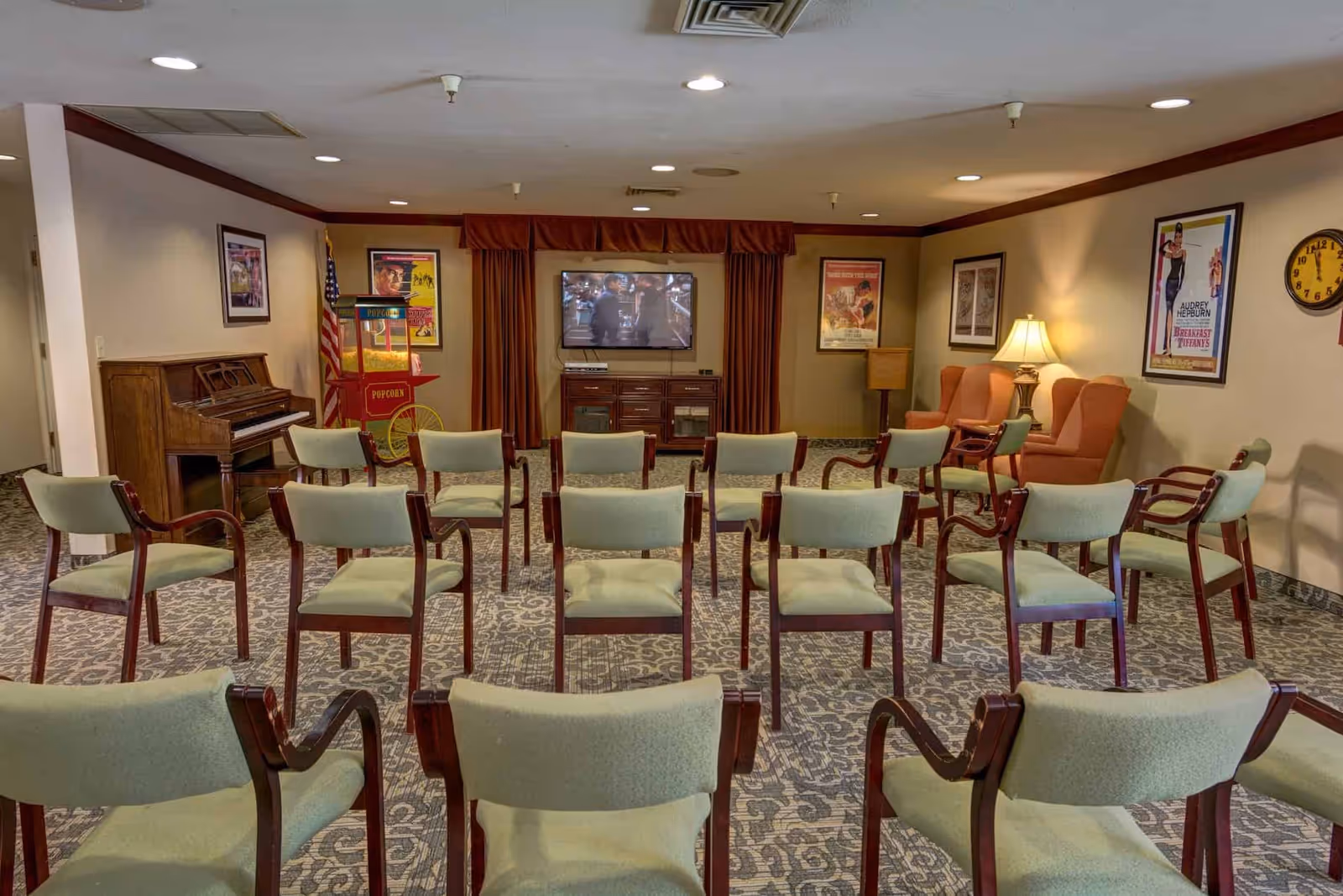 Community seating area with rows of chairs facing a TV, piano, popcorn cart and armchairs in a senior living facility.