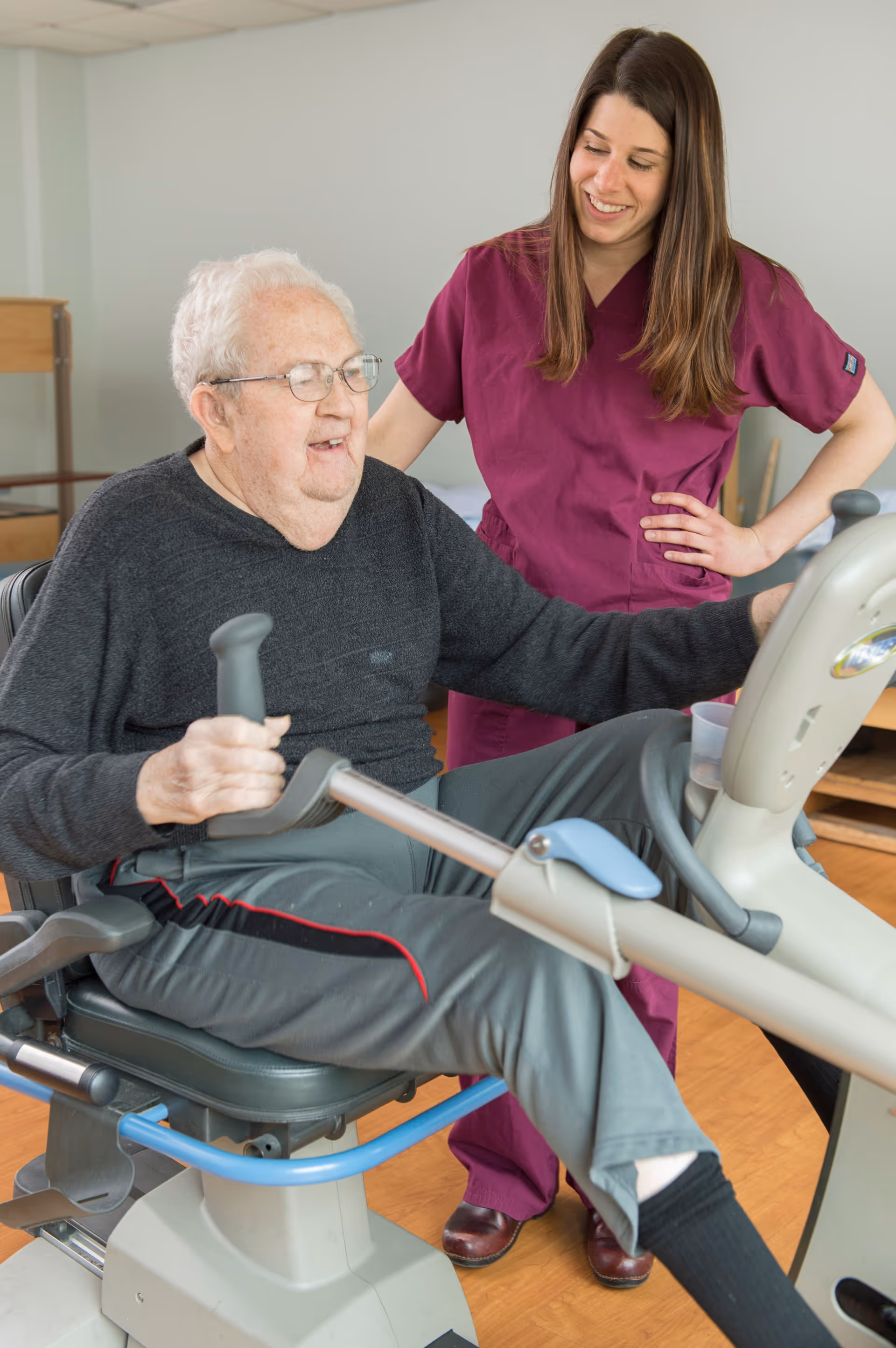 An elderly man with white hair and glasses is exercising on a seated stationary bike while a smiling female caregiver in purple scrubs stands beside him, offering support and encouragement in a well-lit room with wooden flooring.