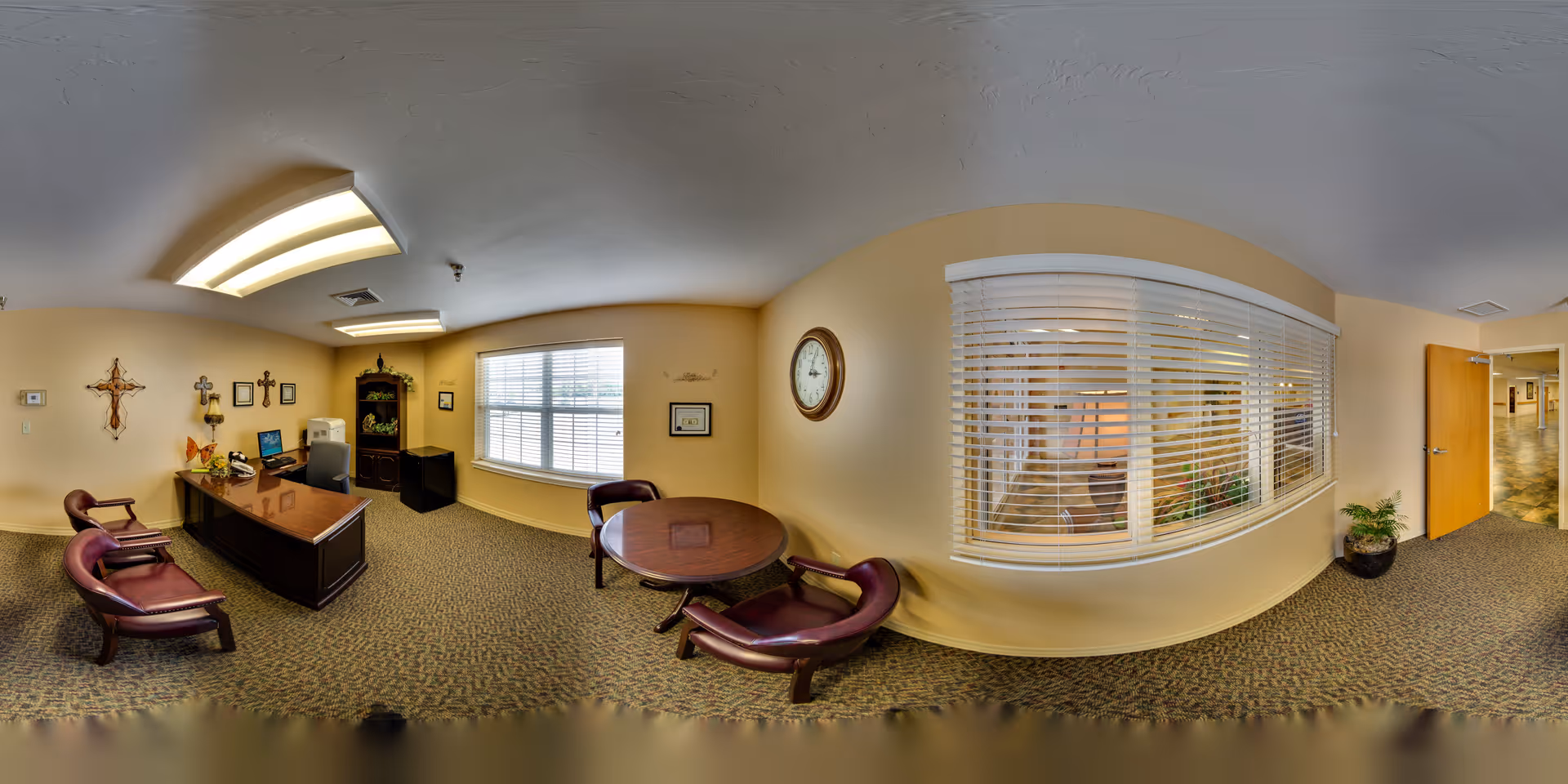 An interior reception/office area with a polished desk, several leather chairs and a round table, a large window with blinds, wall clock, and a hallway entrance.