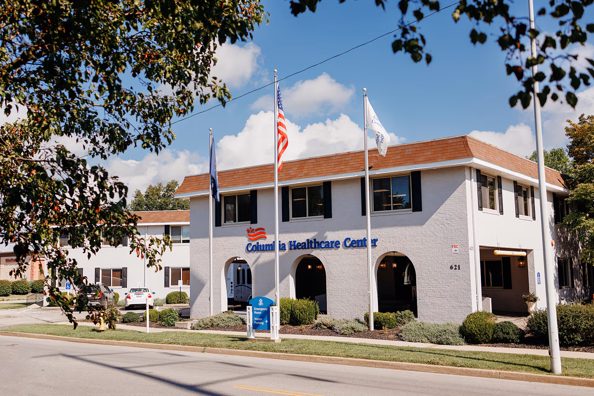 Exterior view of Columbia Healthcare Center, a two-story building with white walls and a brown roof. Three flagpoles stand in front of the building, displaying the American flag and two other flags. There are bushes and small trees around the building, and a blue sign indicating emergency room and visitor parking. A few cars are parked near the building under a clear sky with some clouds.